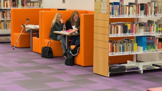 ruimte in een bibliotheek, op afstand zitten studenten in oranje banken te studeren, tussen de boeken in wandrekken