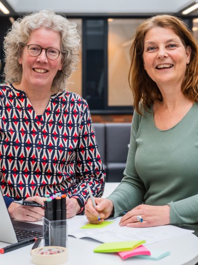 Petra Siemonsma en Arlette Hesselink staan bij elkaar aan een tafel met laptop