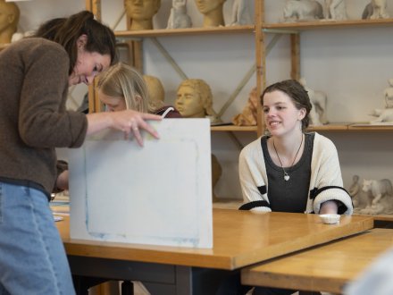 Docent toont een werkstuk aan een student tijdens een les Beeldende Therapie in een atelier met gipsmodellen op de achtergrond.