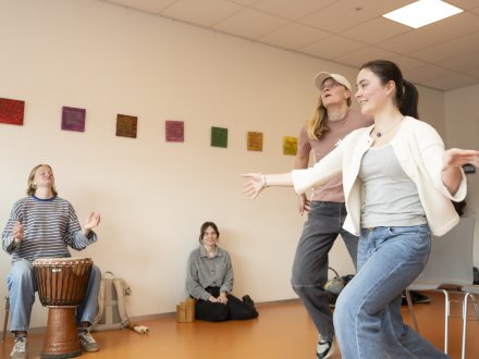Twee studenten Spraak- en dramatherapie oefenen een bewegings- en expressieoefening, terwijl een docent muziek maakt op een djembé en medestudenten toekijken.