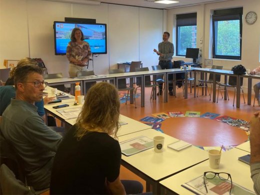 Meerdere mensen zitten in een lokaal aan tafels die in een vierkant staan, en er staat iemand voor een digibord te presenteren. Ook liggen er allemaal gekleurde a4'tjes in een cirkel in het midden van de tafels.