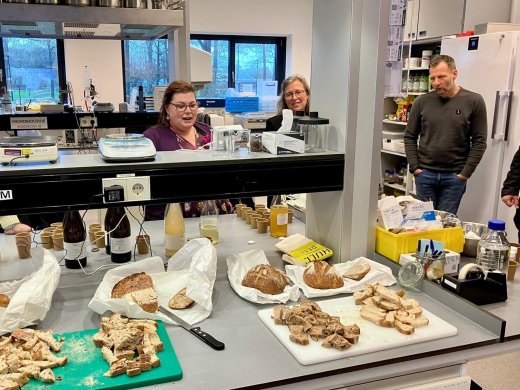 Een aanrecht in een laboratorium. Hierop liggen verschillende soorten brood en Kombucha. Er staan drie mensen omheen.