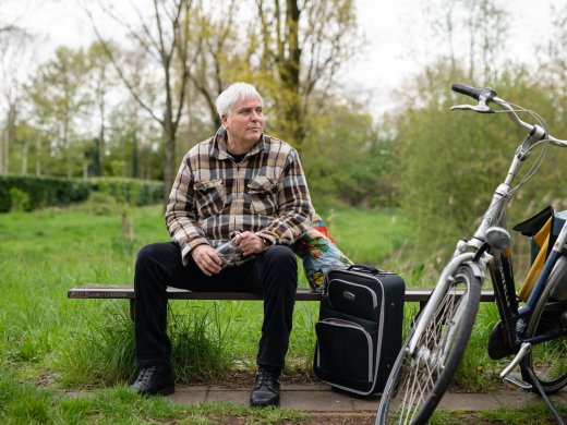 Foto van een man op een bankje met een koffer en fiets ernaast