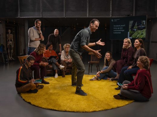 Een man vertelt beeldend en een groep mensen luistert aandachtig en geamuseerd.