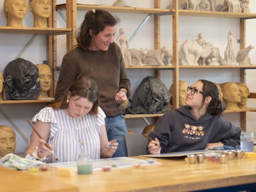 Docent begeleidt twee studenten tijdens een praktijkles Beeldende Therapie in een atelier met beeldhouwwerken en creatieve materialen.