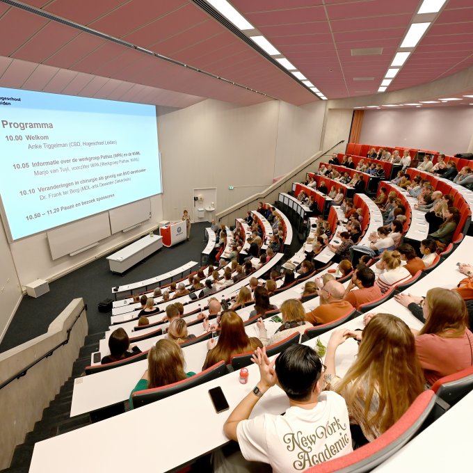 Bezoekers zitten in zaal en kijken naar presentatie waar het programma op te zien is.