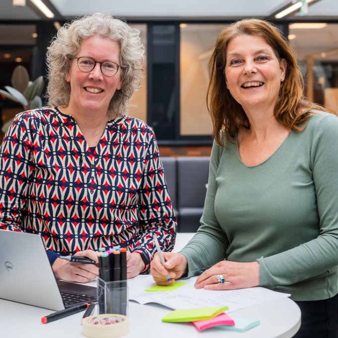 Petra Siemonsma en Arlette Hesselink staan bij elkaar aan een tafel met laptop