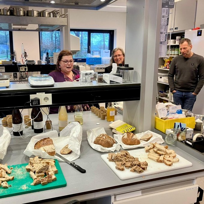 Een aanrecht in een laboratorium. Hierop liggen verschillende soorten brood en Kombucha. Er staan drie mensen omheen.
