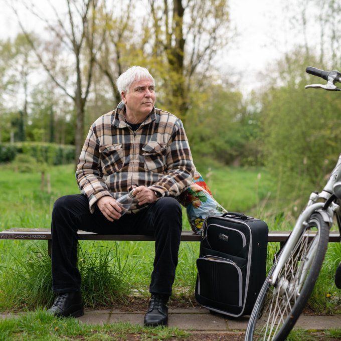 Foto van een man op een bankje met een koffer en fiets ernaast